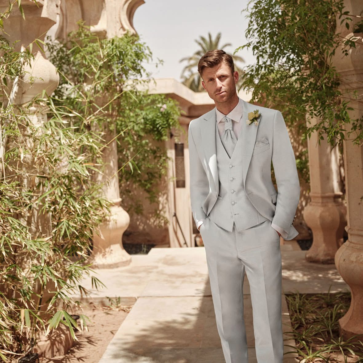 Ein Mann in einem hellblauen Anzug steht selbstbewusst in einem sonnendurchfluteten, von Grün umgebenen Innenhof.