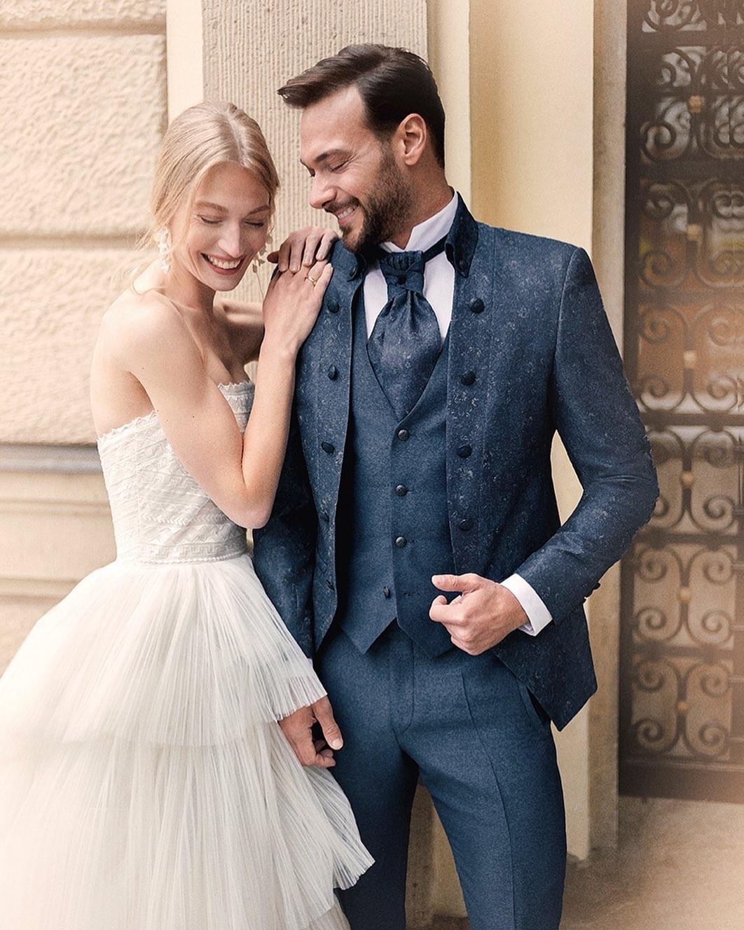 Ein Mann und eine Frau in eleganter Hochzeitskleidung stehen nebeneinander und lächeln in die Kamera in einer romantischen Kulisse im Freien.